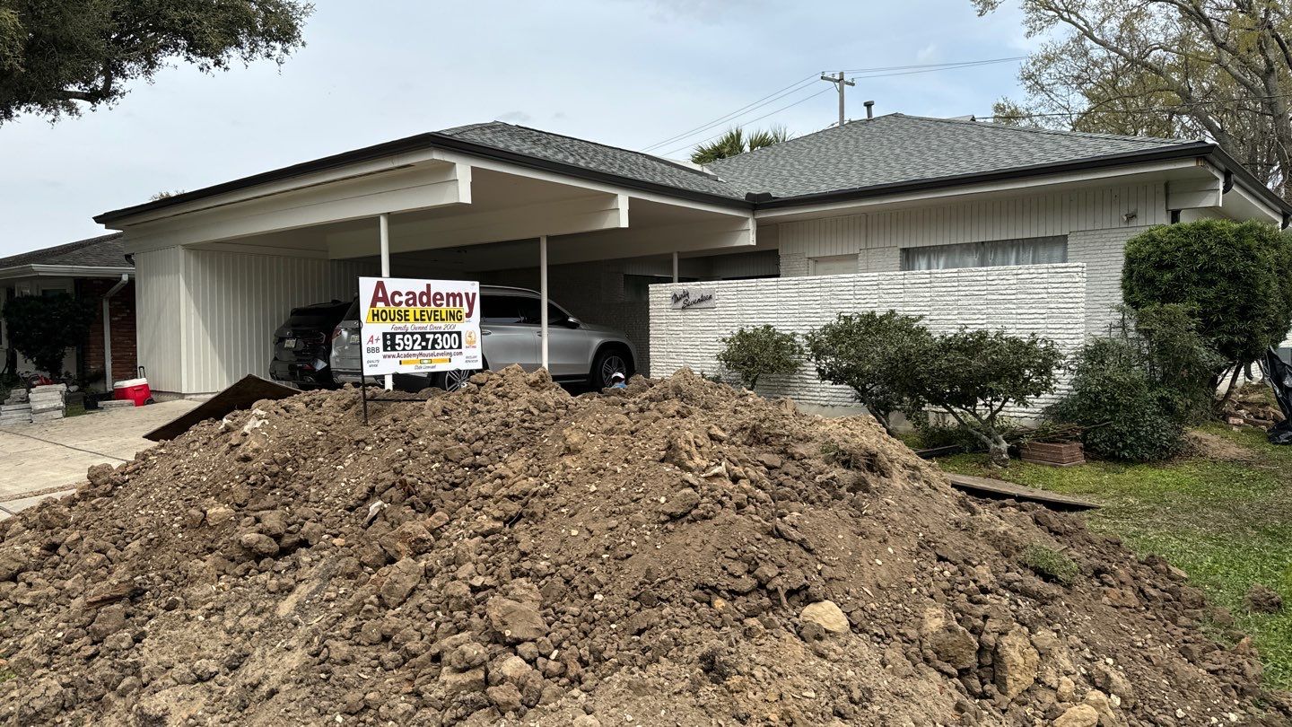 A large pile of dirt is situated in the driveway of a residential home in the Southeast, with an "academy house leveling" sign posted, indicating foundation repair methods or leveling work in progress.