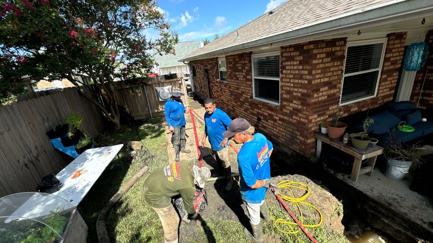 Five workers in a backyard with gardening tools and hoses, engaged in a house leveling project near a red brick house. There's a wooden fence, some plants, and a blue outdoor couch visible in the scene.