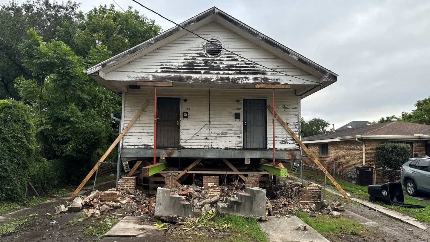 A small house in Jefferson Parish, elevated on wooden beams, shows wear and damage, with bricks scattered around its base and supports steadied on either side. House leveling might be needed to restore stability to this Metairie home.