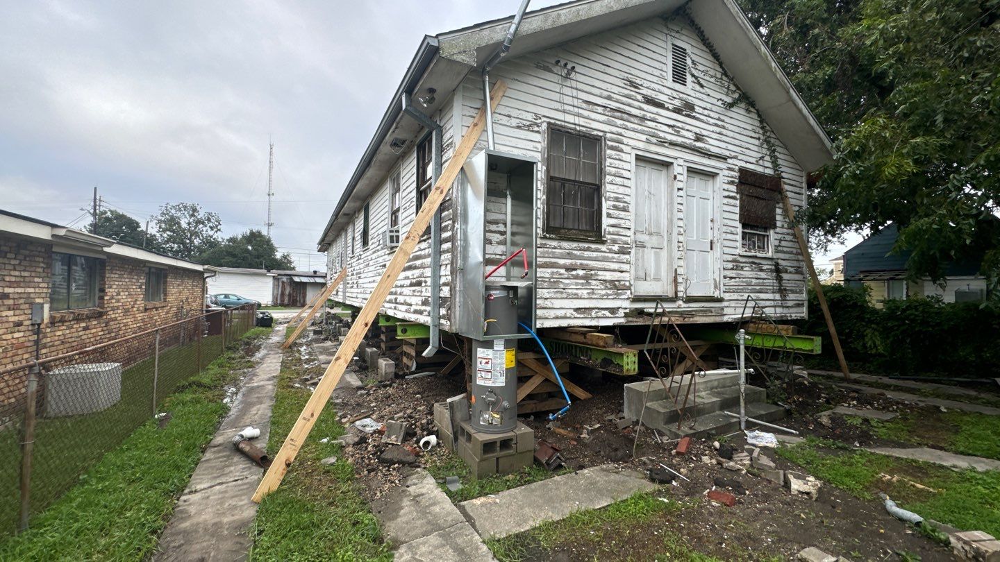 A weathered house in Metairie is elevated on stilts for foundation repair. A hydraulic jack and wooden support beam are visible. Nearby, a fenced yard and the surrounding neighborhood of Jefferson Parish can be seen.