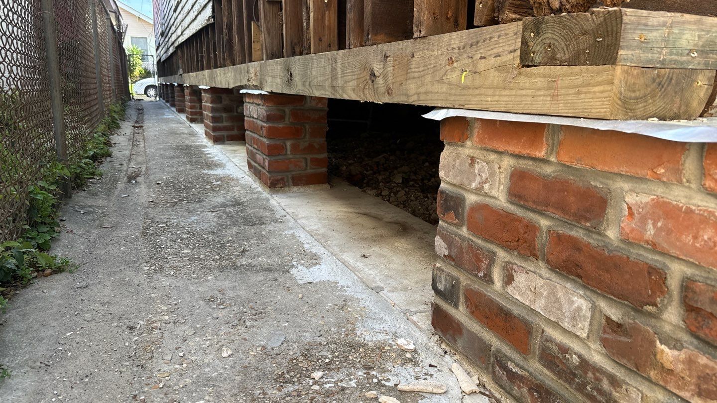 A building foundation supported by brick pillars, with a concrete pathway and metal fence on the left side, showcases the classic techniques of home elevation commonly seen in New Orleans.