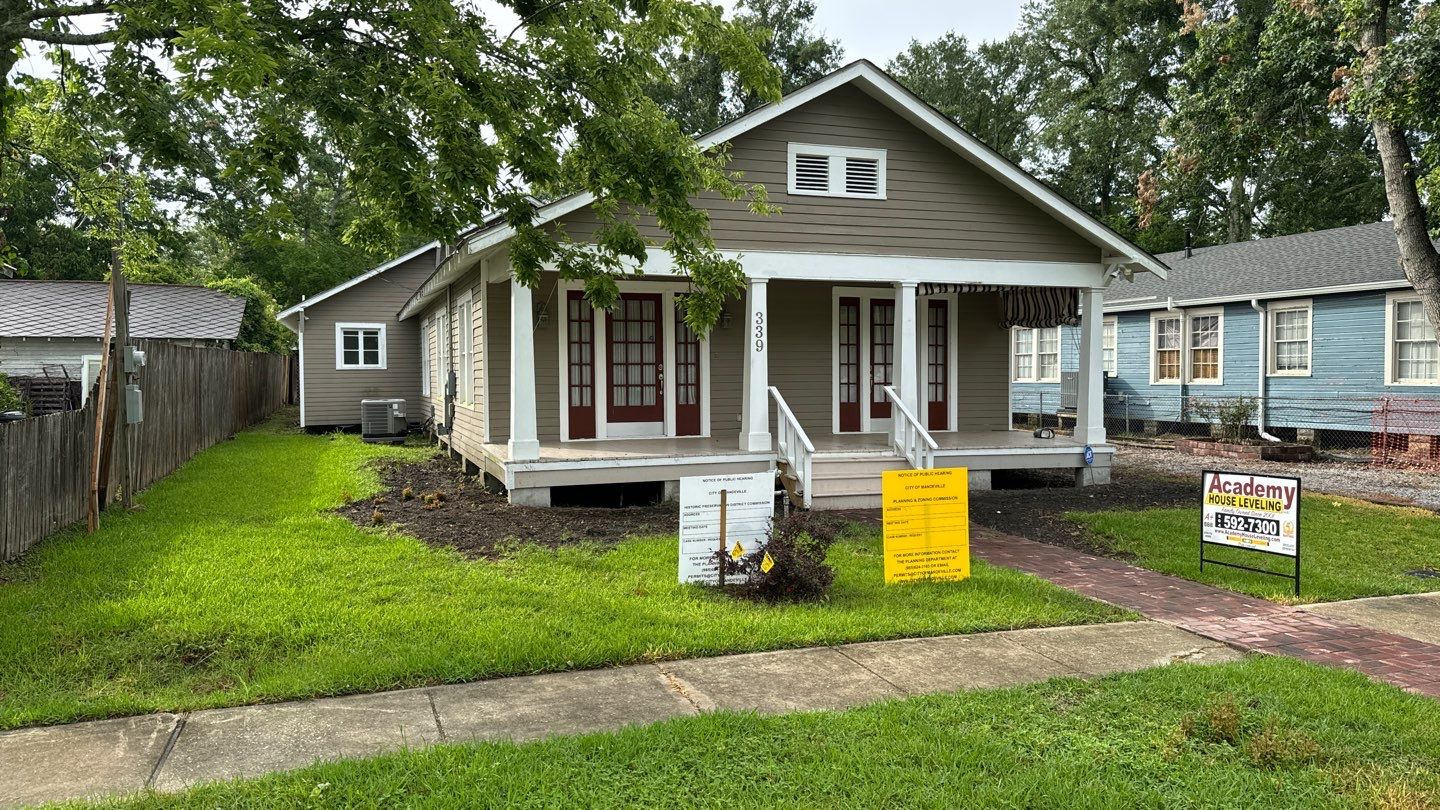 A one-story house with a front porch and steps, beige siding, and white accents. Multiple signs are in the front yard, including for sale and construction signs, indicating possible foundation repair. Neighboring houses are visible.