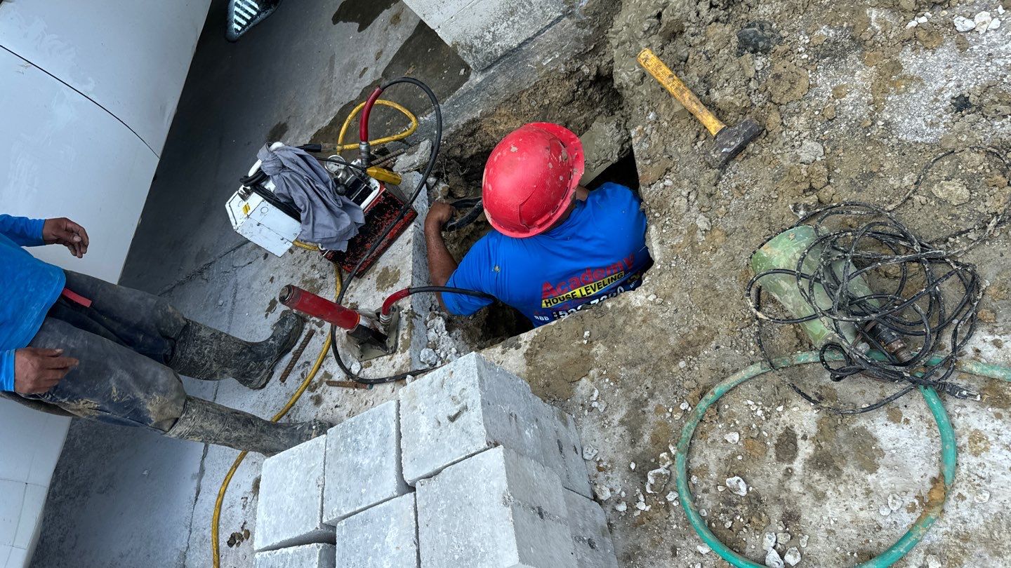 A construction worker wearing a red helmet works on the foundations inside a hole at a Kenner LA construction site, surrounded by tools, equipment, and bricks. Another worker stands nearby, ready to address any drainage issues that may arise.
