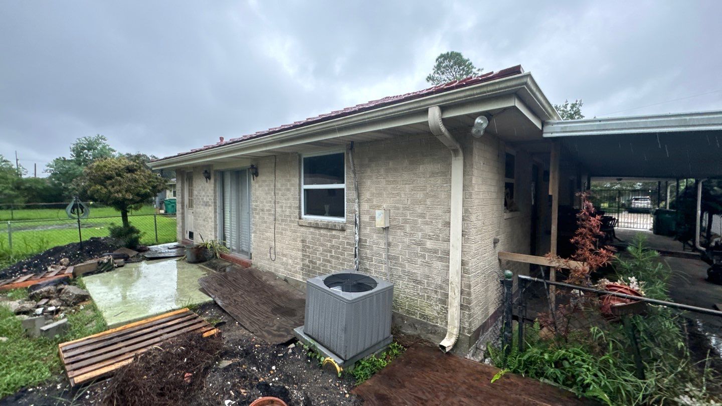 A small beige brick house in Louisiana, featuring a red roof and a metal awning, stands on a concrete patio surrounded by a wet yard. An air conditioning unit is nestled next to the home. The overcast sky adds to the charm of this level-a-home beauty.