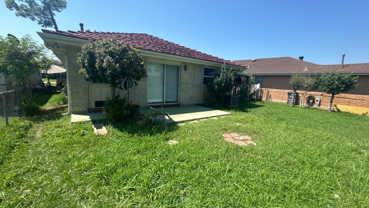 A small backyard with a grassy lawn, concrete patio, and potted plants next to a single-story house that recently underwent home leveling in Louisiana, featuring a charming tiled roof.
