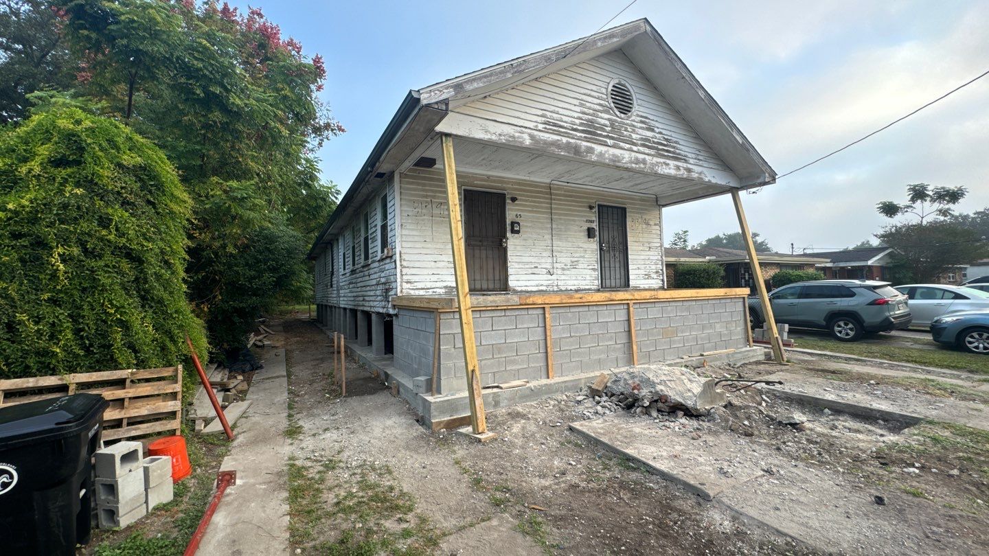 A single-story white house in New Orleans, set on a raised foundation, is undergoing home leveling and renovation. Piles of debris are scattered in front, with a couple of parked cars nearby.
