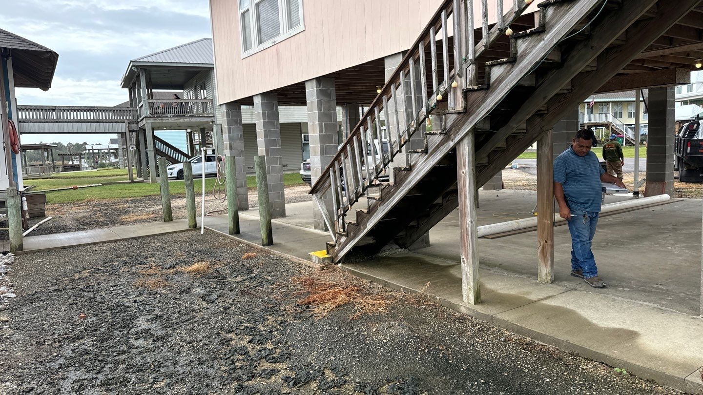 A man stands under a raised wooden house by a staircase, reminiscent of the homes in New Orleans. Nearby, there's a muddy area and another elevated house with a walkway. The cloudy sky looms ahead, illustrating the challenges of leveling a pier and beam foundation in this climate.