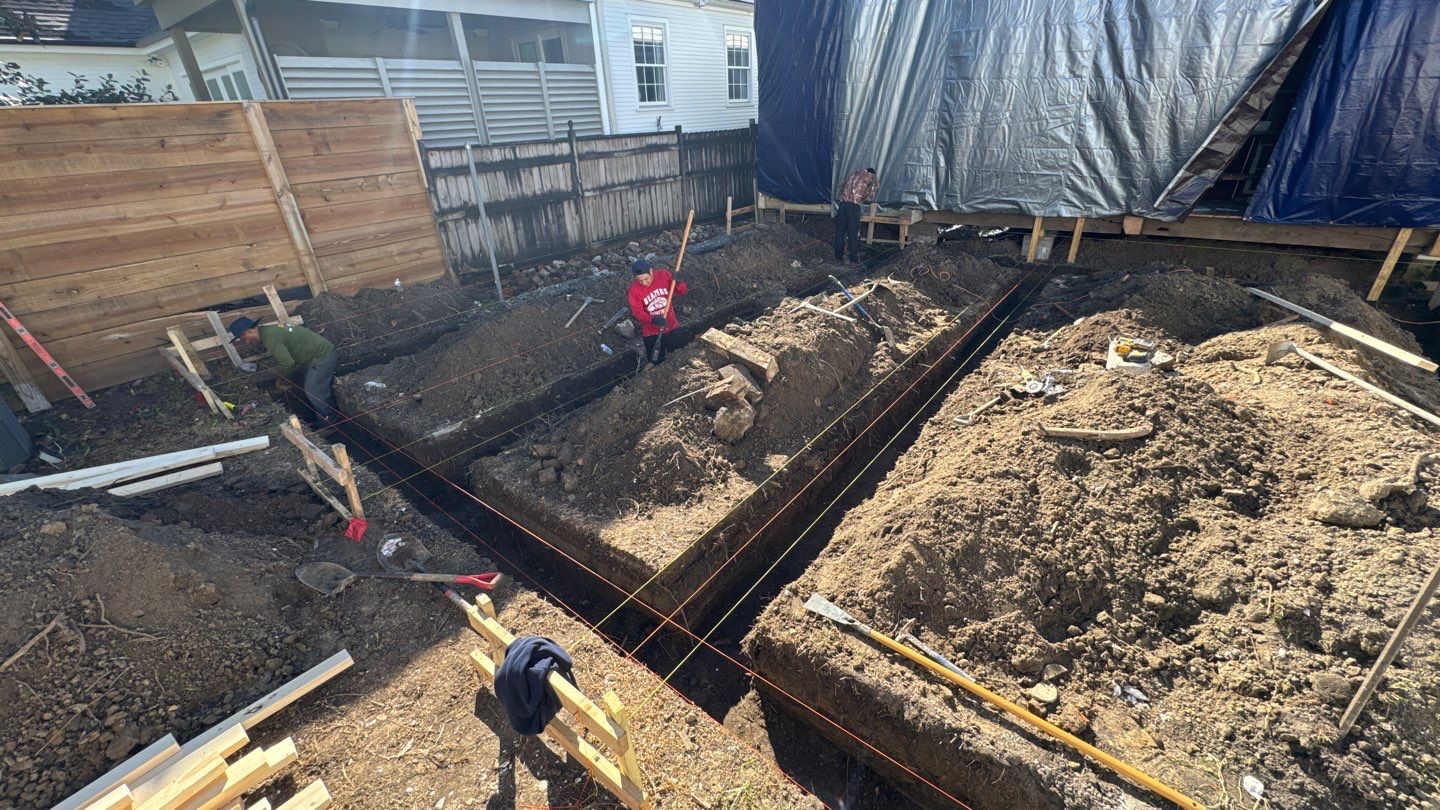 Two construction workers are diligently digging trenches for foundations at a Louisiana residential site. The area is marked with red string lines and surrounded by piles of dirt, while nearby, a building is covered with tarps to prevent any weather-related fail.